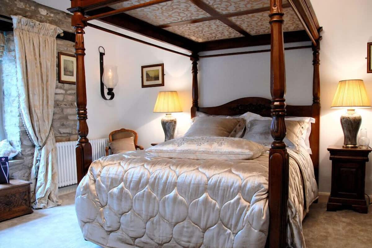 Uma cama de dossel com cabeceira acolchoada, duas mesas de cabeceira com abajures, uma parede de pedra e um radiador.