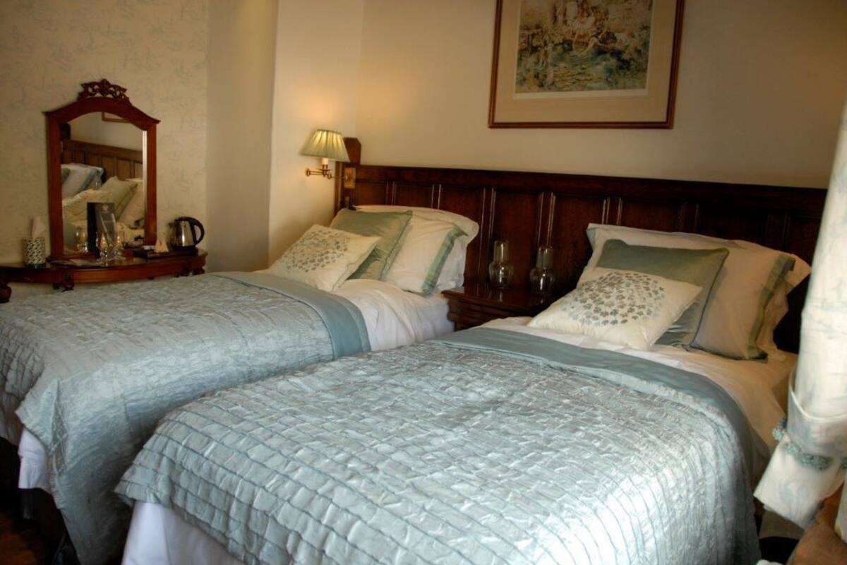 Quarto de hotel com duas camas, cabeceira de madeira, mesa de cabeceira com abajur e um quadro na parede.