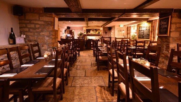 A restaurant with stone walls, wooden tables and chairs, and a bar area. - Busfeild Arms (Keighley)