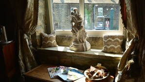 A room with a window seat featuring a statue, a table with magazines and a bowl of food, and a view of a brick building outside. - Busfeild Arms (Keighley)