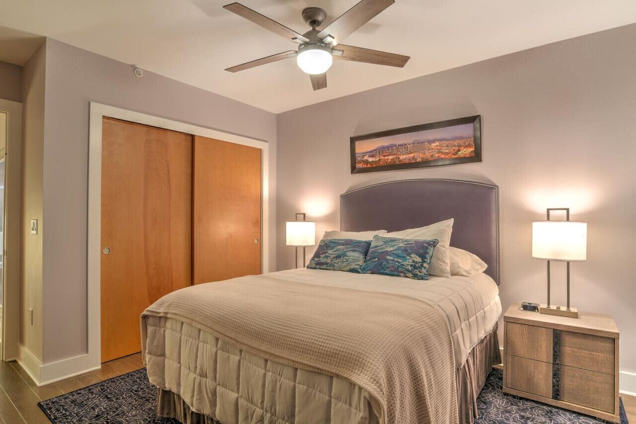 A bedroom with a bed, two bedside lamps, a ceiling fan, a cityscape painting, and a wooden wardrobe.