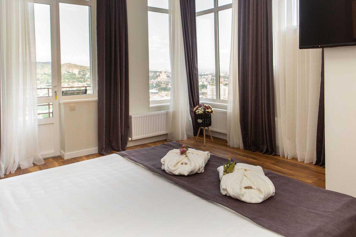 Un cuarto de hotel con una cama grande, vistas de la ciudad y una mesita con un florero.