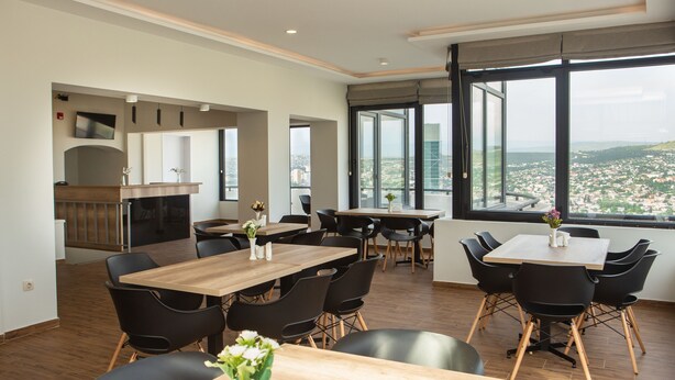 Un comedor moderno con mesas de madera y sillas negras, ventanas amplias con vista a la ciudad, y una zona de cocina al fondo.