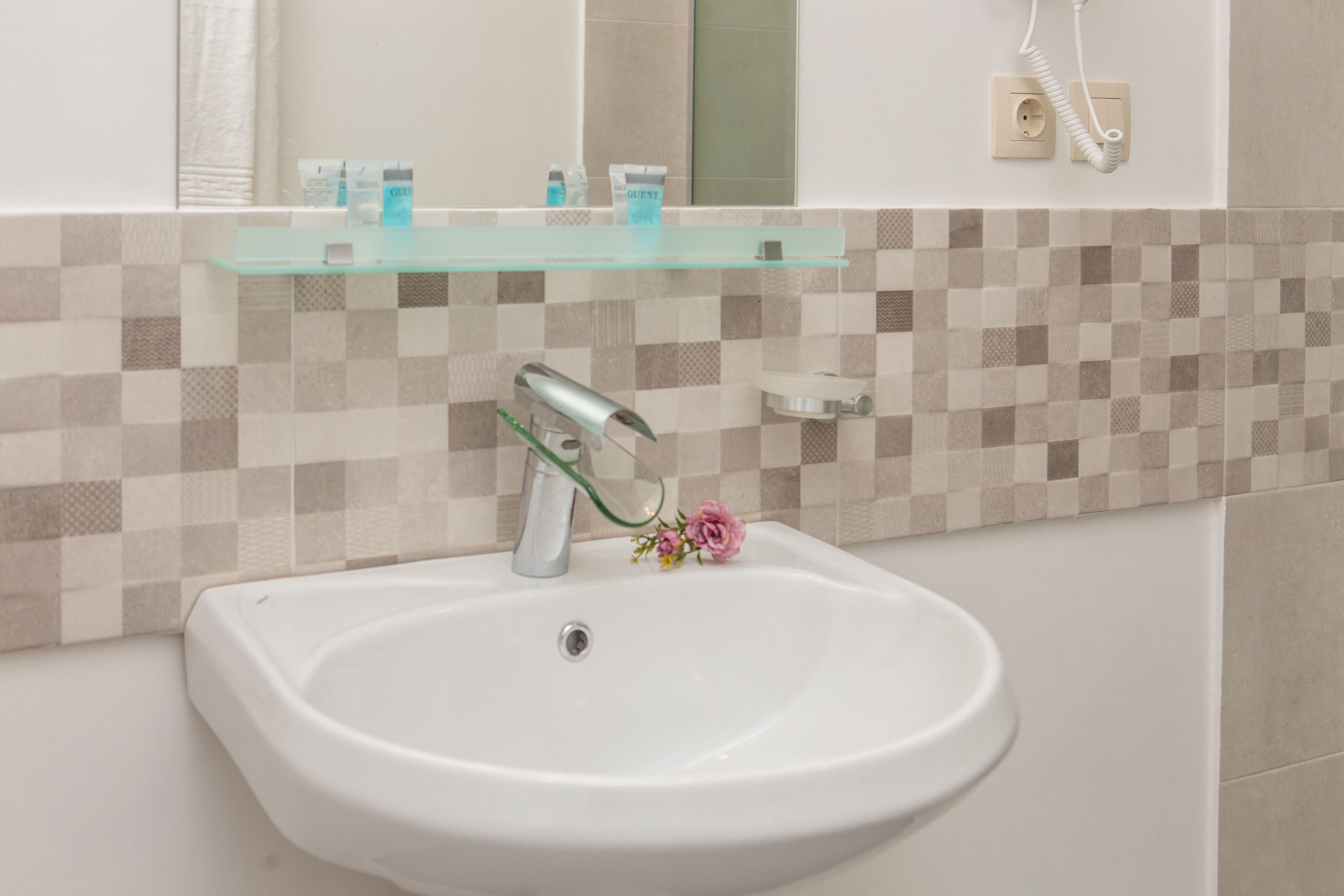 A bathroom with a white sink, a glass shelf above it, and a tiled wall.