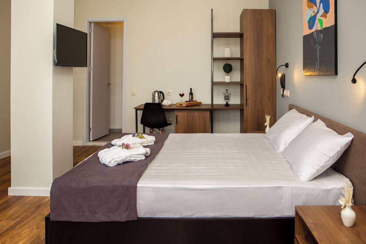 Una cama bien hecha con ropa blanca, un escritorio con una silla, un televisor montado en la pared y un estante con objetos decorativos.
