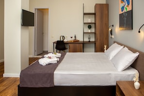 A neatly made bed with white linens, a desk with a chair, a television mounted on the wall, and a shelf with decorative items.