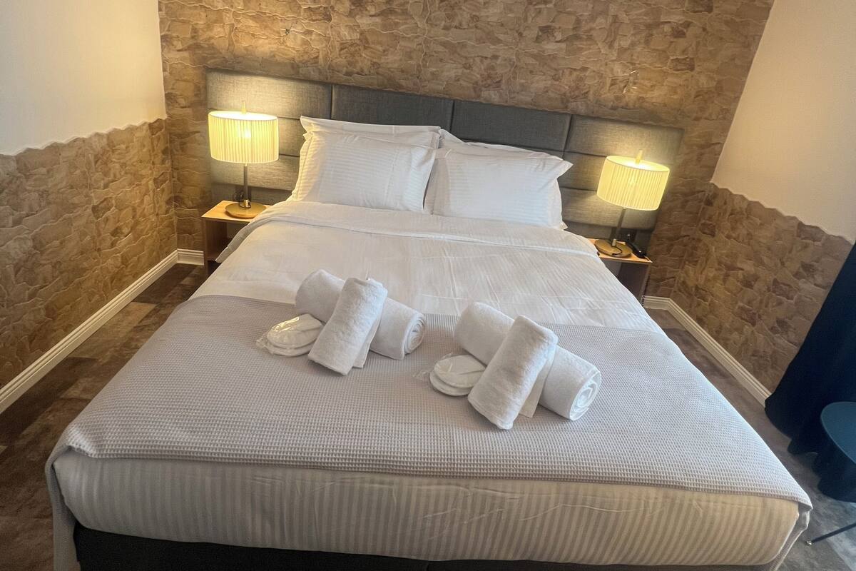 A neatly made bed with white linens and towels, a stone-textured wall behind, and two bedside lamps.