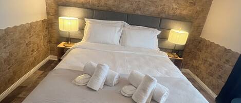 Un lit bien fait, avec du linge de lit et des serviettes blanches, un mur en pierre texturée derrière, et deux lampes de chevet.