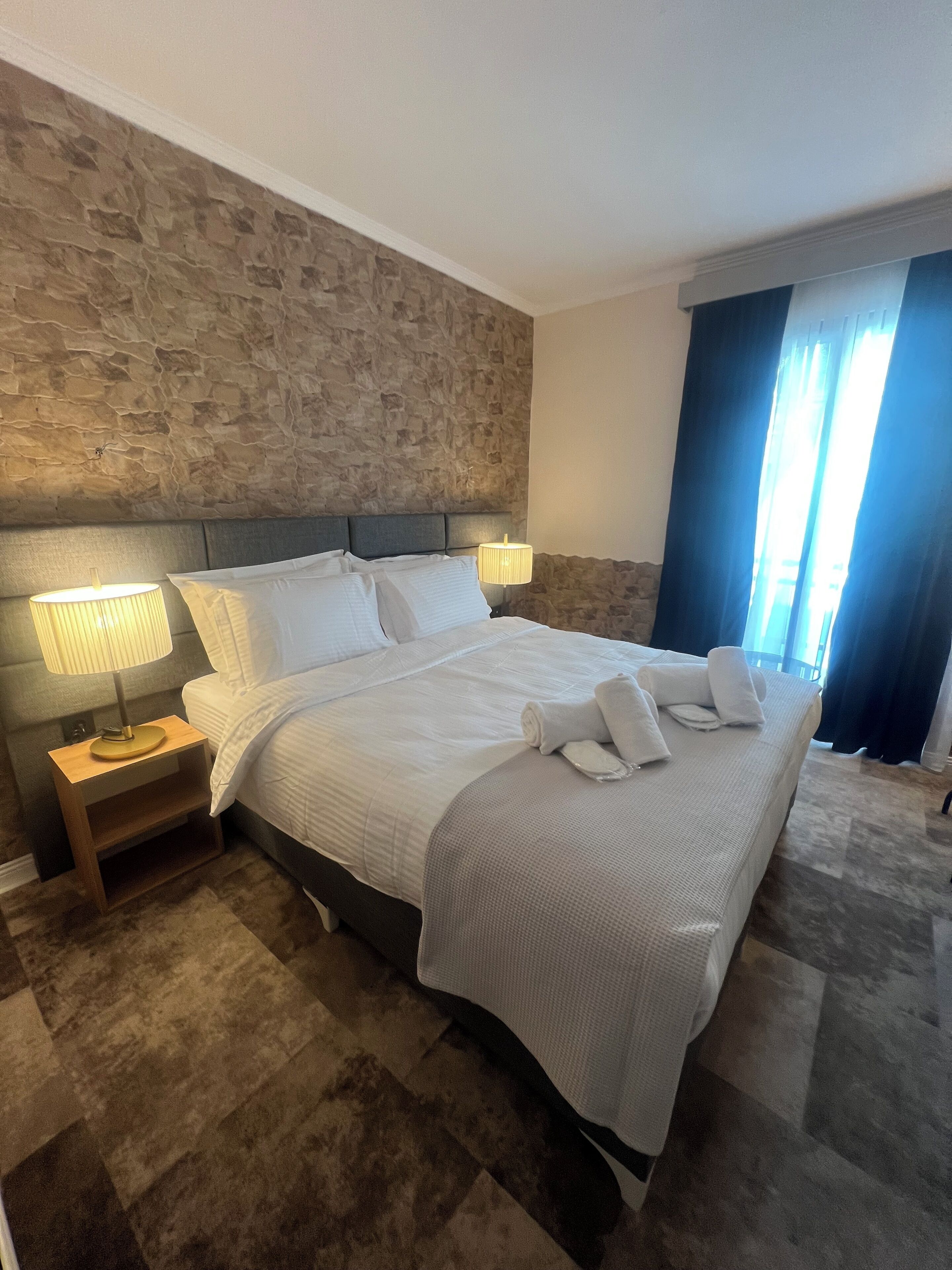 A neatly made bed with white linens, two bedside lamps, and a stone-textured wall behind it.