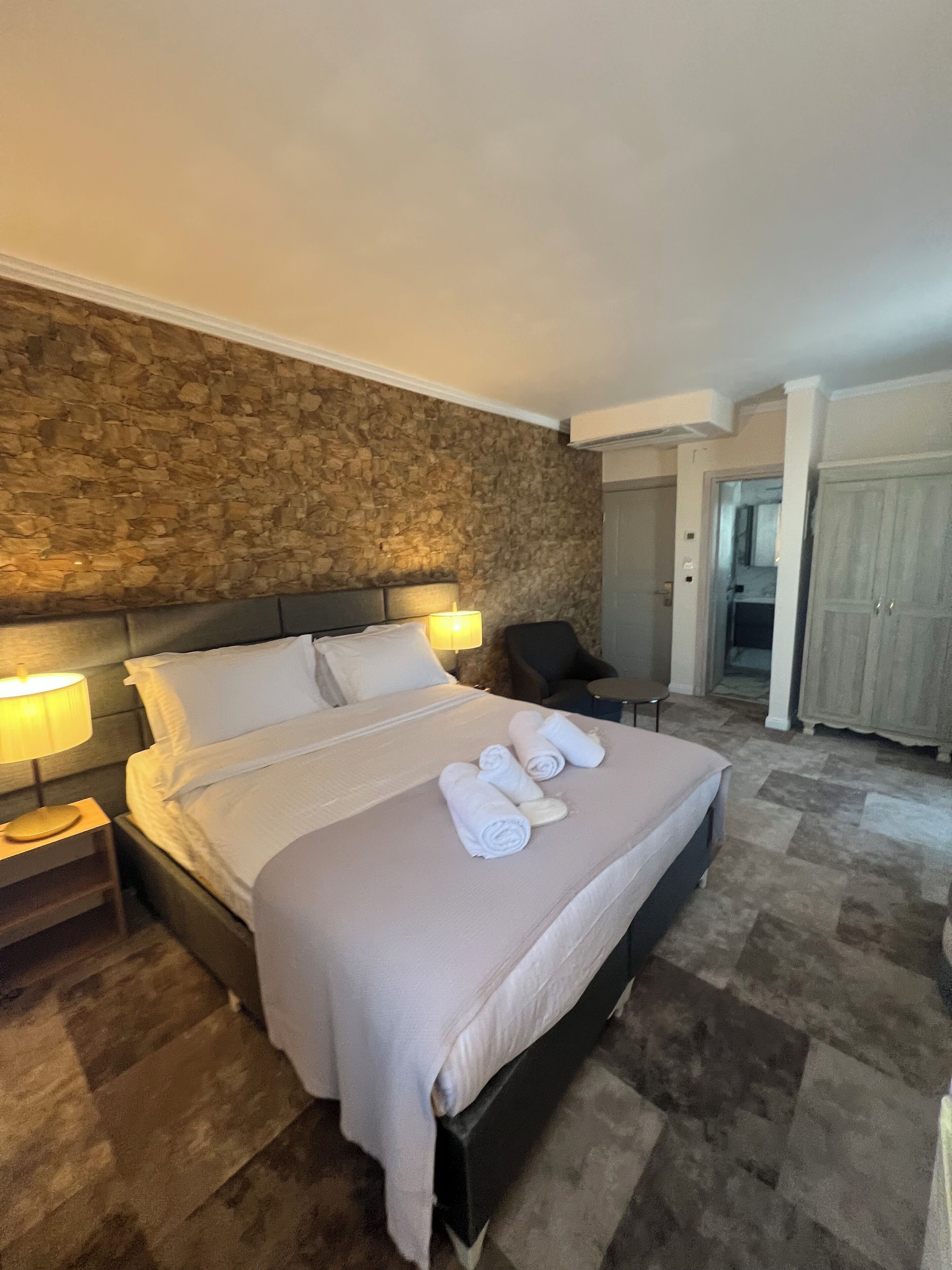 A neatly made bed with white linens and towels, a stone wall behind, a nightstand with a lamp, and a chair in the corner.