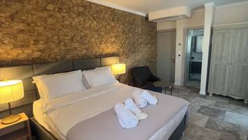 A neatly made bed with white linens and towels, a stone wall behind, a nightstand with a lamp, and a chair in the corner.
