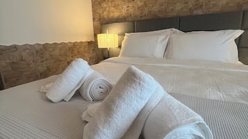 A neatly made bed with white linens and rolled towels, a stone-textured headboard, and a wall-mounted lamp.