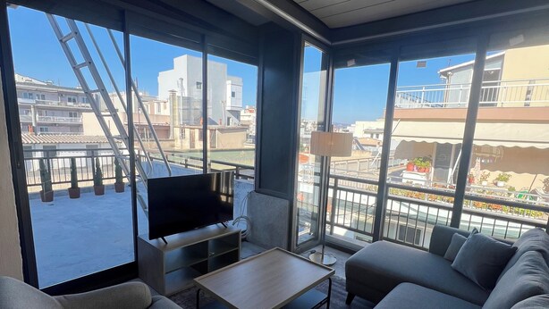 Una sala de estar moderna con sofá, sillones, una mesa de centro y televisor. Amplias ventanas ofrecen vistas panorámicas de la ciudad.