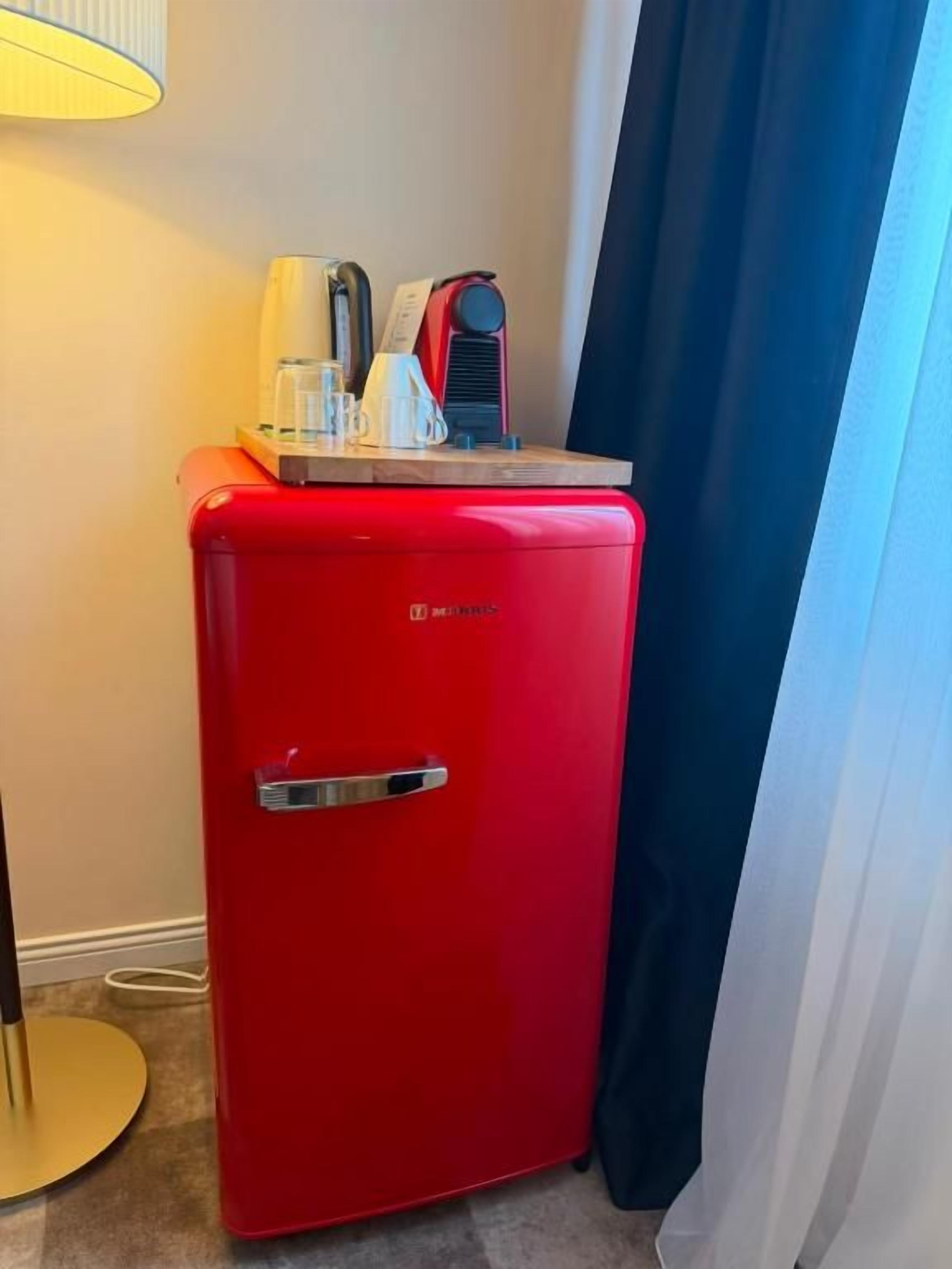 Un refrigerador rojo compacto con asa, colocado junto a una lámpara y una cortina.