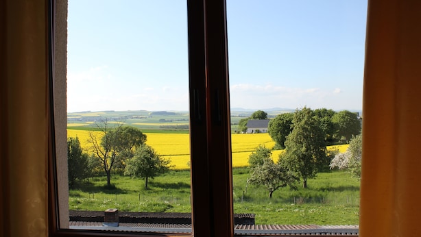 Une vue par la fenêtre sur un paysage rural, avec des champs, des arbres et une maison au loin.