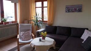 Un salon comprenant un canapé, un fauteuil, une table avec un vase de fleurs, une plante en pot et une fenêtre avec des rideaux.