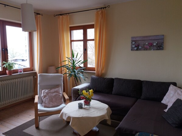 A living room with a sofa, a chair, a table with a vase of flowers, a potted plant, and a window with curtains. - Ferienwohnung zur Burg Eltz (Wierschem)
