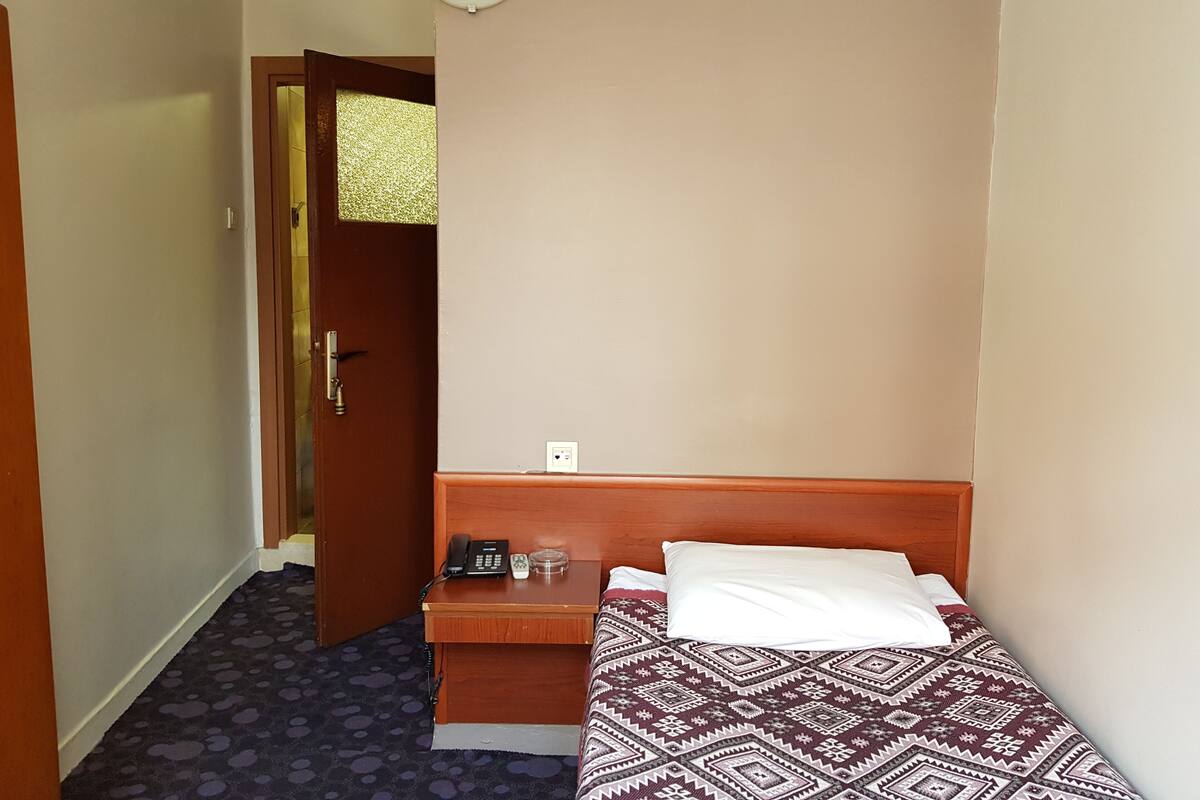 Pequeña habitación individual con una cama y colcha estampada, una mesita de noche con un teléfono y un vaso, y una puerta que da a otra habitación.