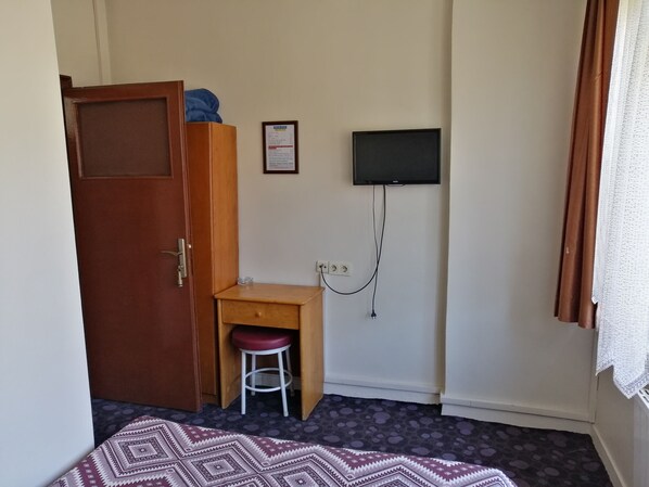 Una habitación con un armario de madera, un escritorio pequeño con un taburete, un televisor montado en la pared y una alfombra estampada en el suelo.