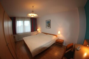 A bedroom with a bed, bedside lamps, a radiator, a window with curtains, and a picture on the wall.