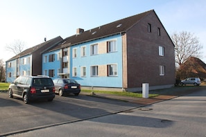 Un edificio residencial de dos pisos con fachada azul y tejado marrón. Hay dos coches estacionados adelante.