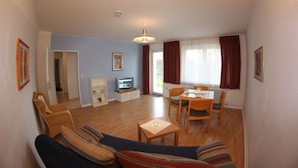 A living room with a sofa, a dining table with chairs, a television, and a window with curtains. - Haus Marinus (Fehmarn)
