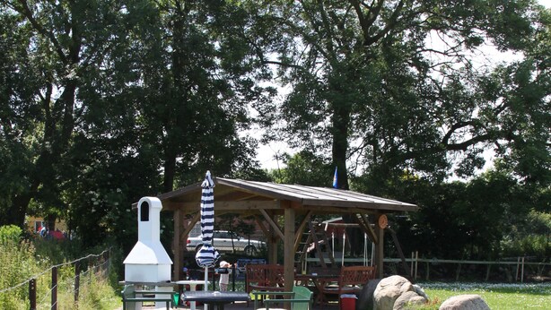 An outdoor seating area with a wooden table and chairs, a white barbecue grill, and a covered pavilion. - Haus Marinus (Fehmarn)