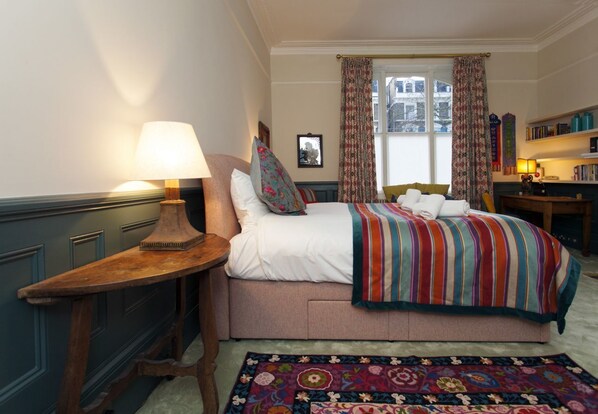 A bedroom with a bed, a nightstand, a lamp, a window with curtains, a bookshelf, and a floral rug.