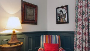 A room with a wooden side table, a lamp, a striped armchair with a red pillow, and patterned curtains.