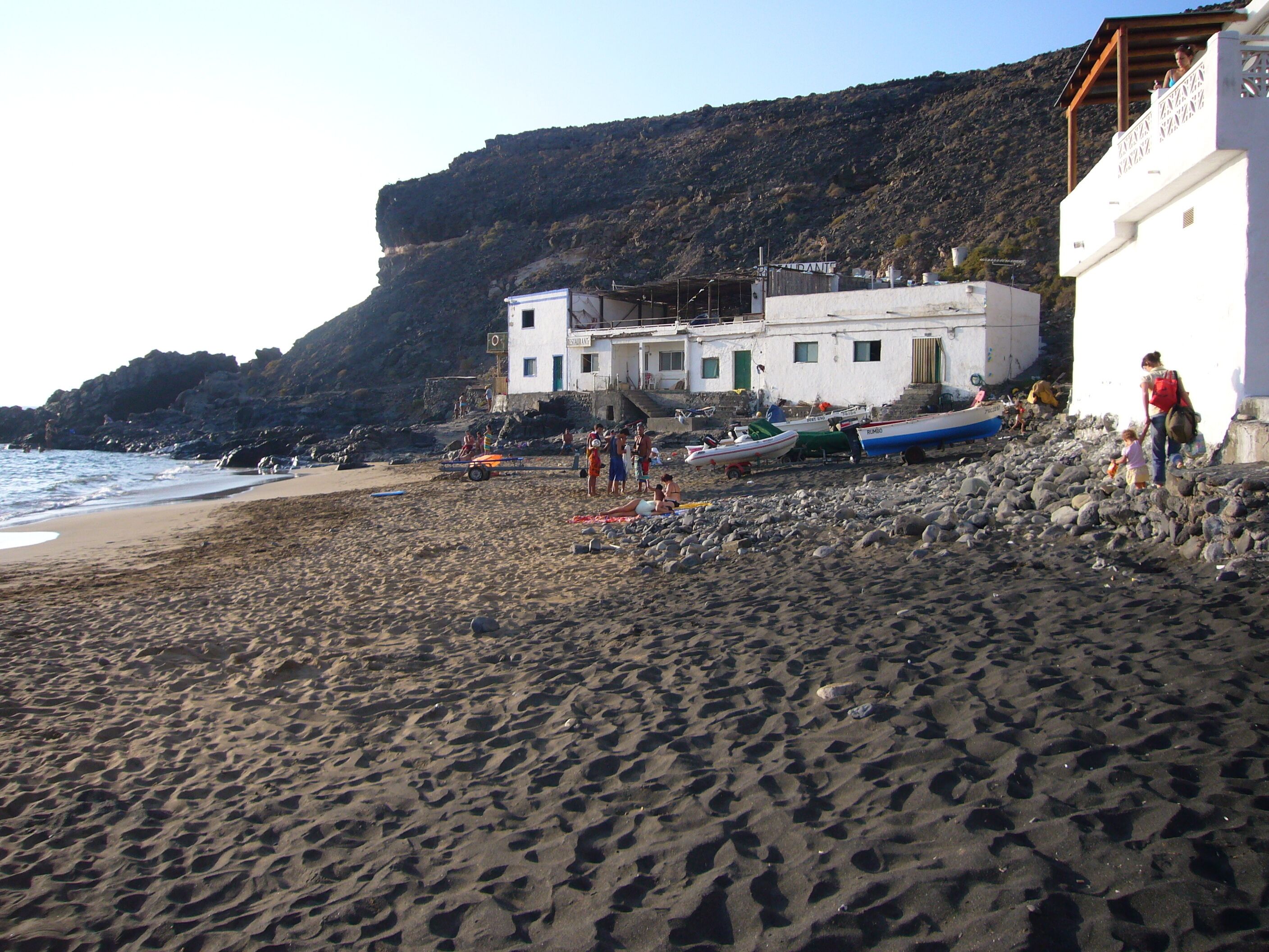 A beach with rocky terrain, a building with a restaurant sign, and boats.