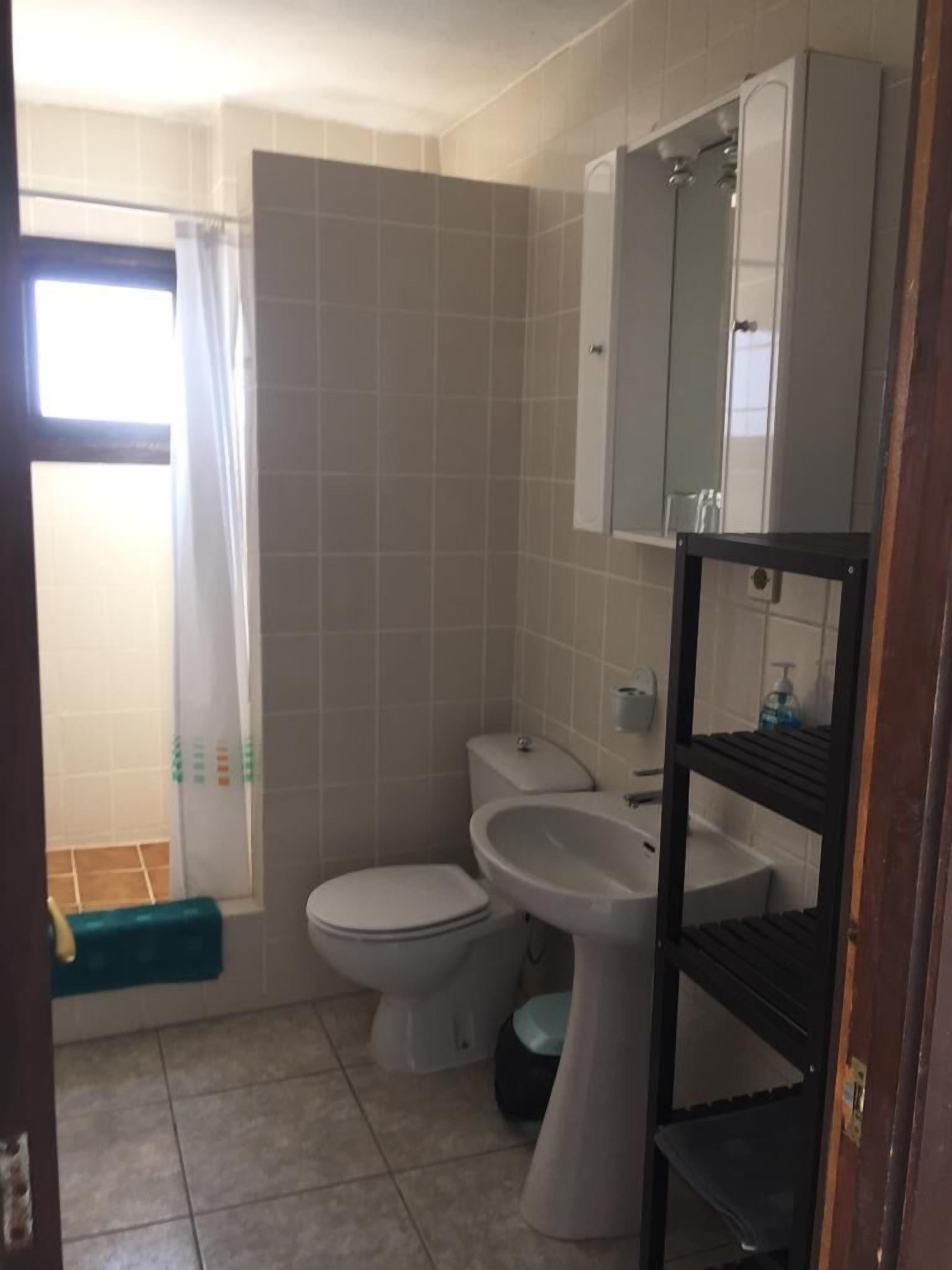 A small, clean bathroom with a toilet, sink, and mirror. There is a shower curtain and a shelf with toiletries.