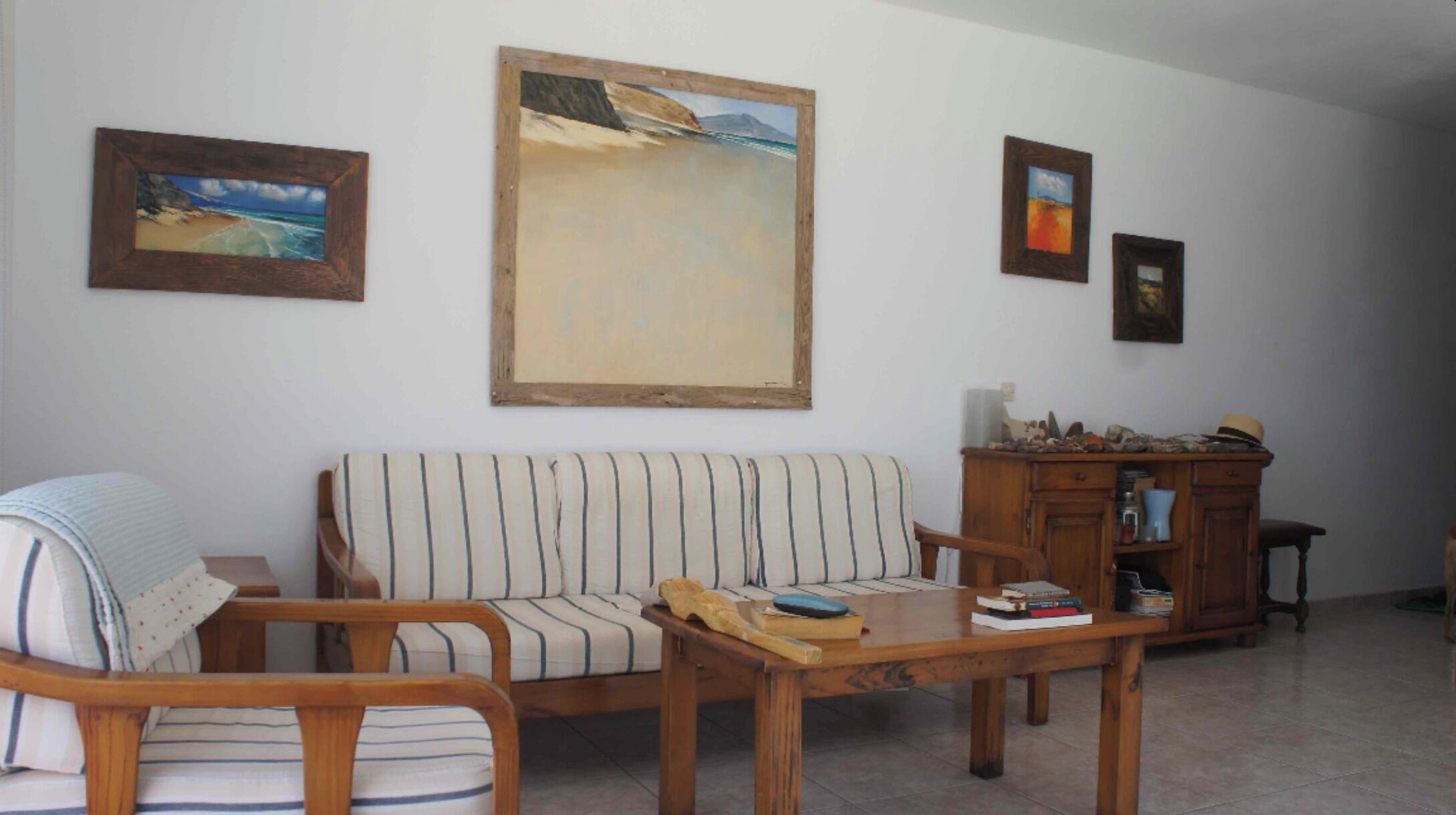 A living room with a striped sofa, a wooden coffee table, and a cabinet with decorative items. There are three framed paintings on the wall.