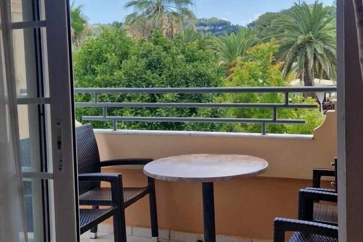 A balcony with a table and chairs, a view of greenery and palm trees, and a clear sky.