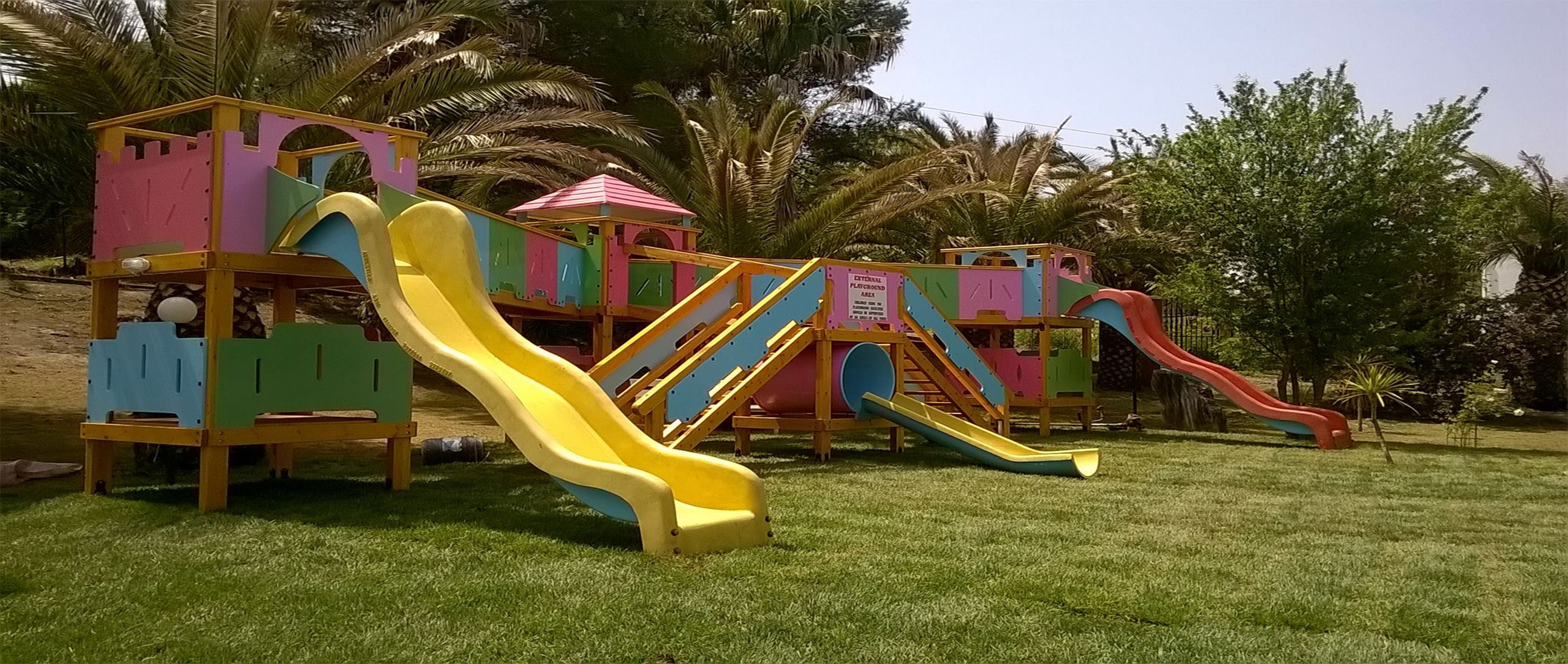 Une aire de jeux colorée, dotée de nombreux toboggans et de structures d’escalade, entourée de palmiers et d’herbe.