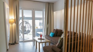 Una sala moderna con un sofá marrón, una mesita con una botella y un vaso, una lámpara de pie y vistas a un espacio exterior con una silla colgante y una mesa.