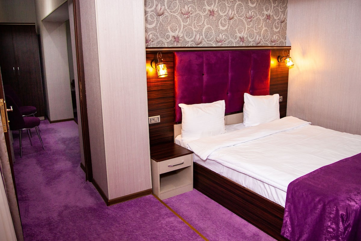 A hotel room with a bed, a nightstand, a chair, and a wall with a floral pattern.