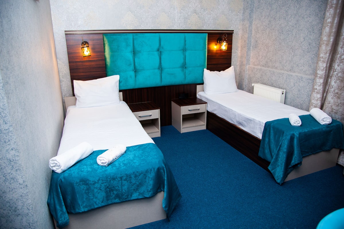 A hotel room with two beds, each with a teal bedspread and white linens. There are two bedside tables with lamps, a wooden headboard, and a blue carpet.
