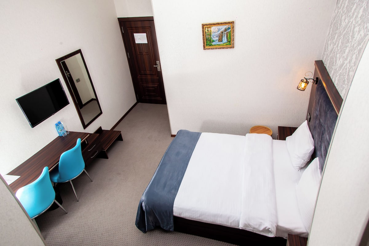 A hotel room with a bed, a desk with chairs, a TV, a door, and a painting on the wall.