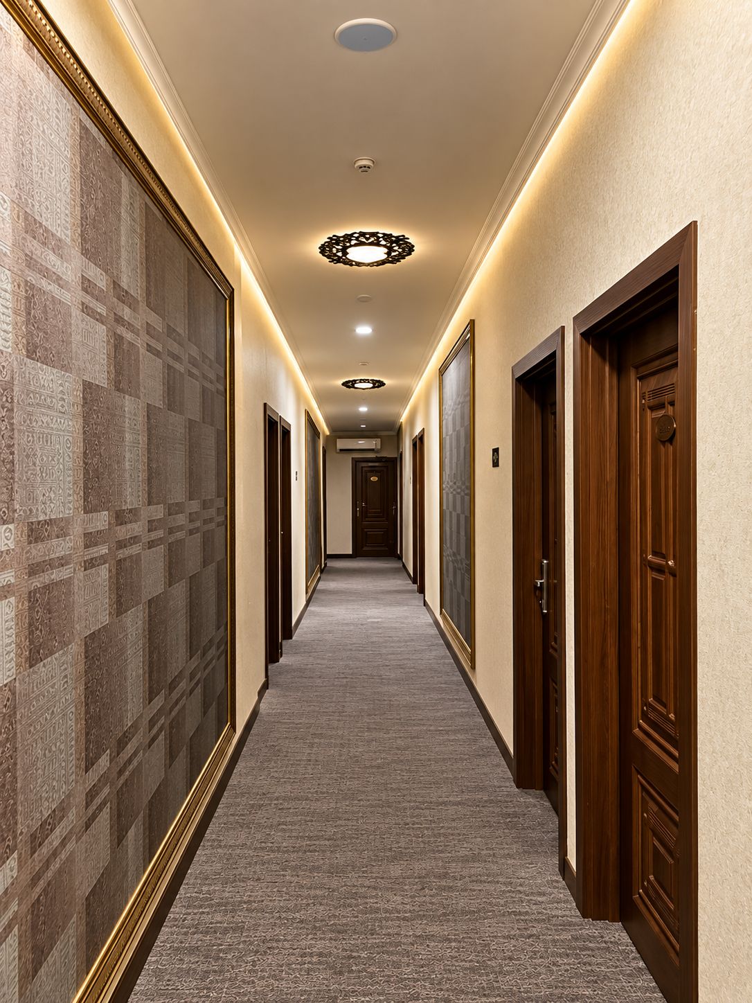 A hotel hallway with carpeted floors, wooden doors, and patterned wall tiles.