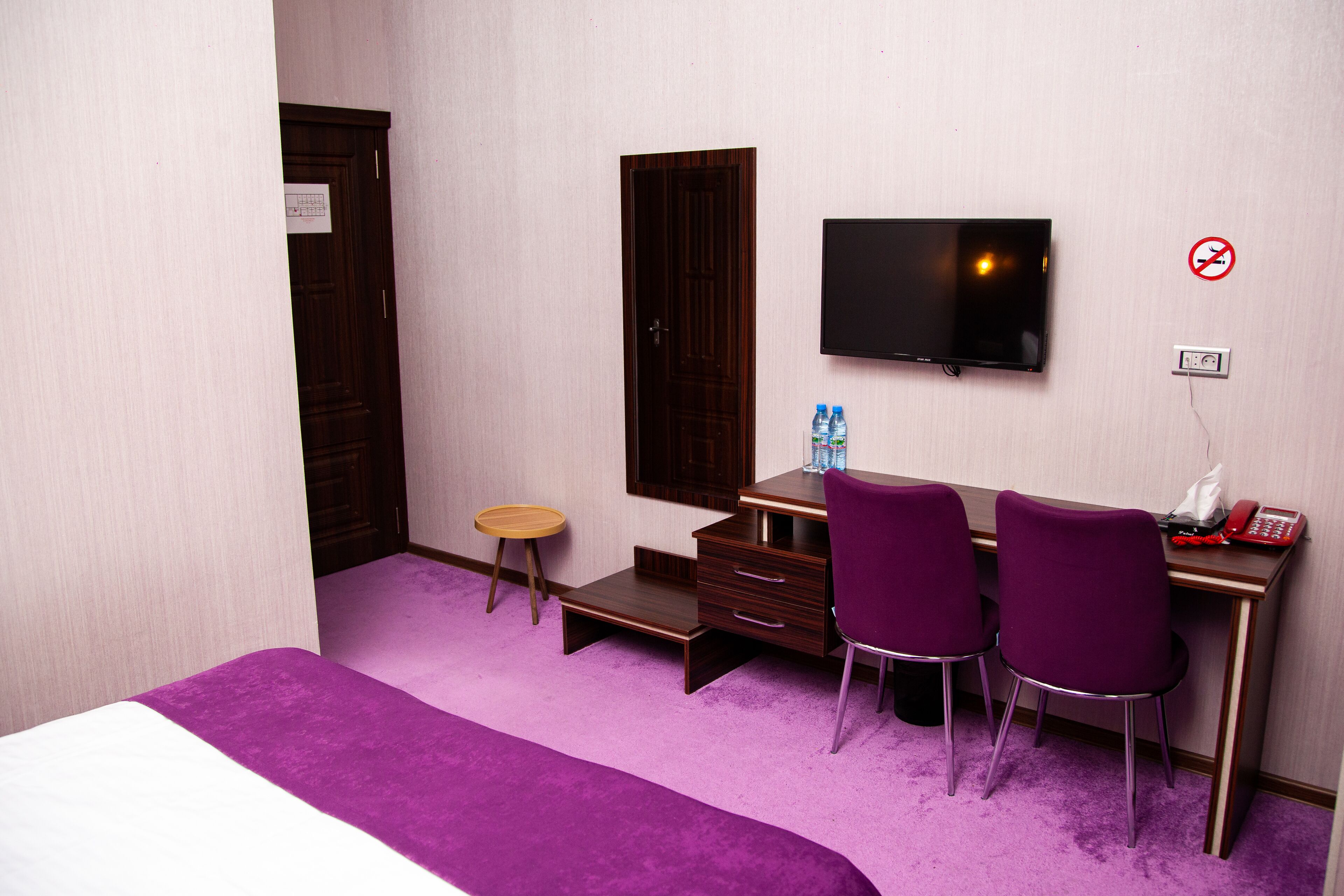 Habitación de hotel con cama, escritorio, dos sillas, televisión, un taburete pequeño y una botella de agua.