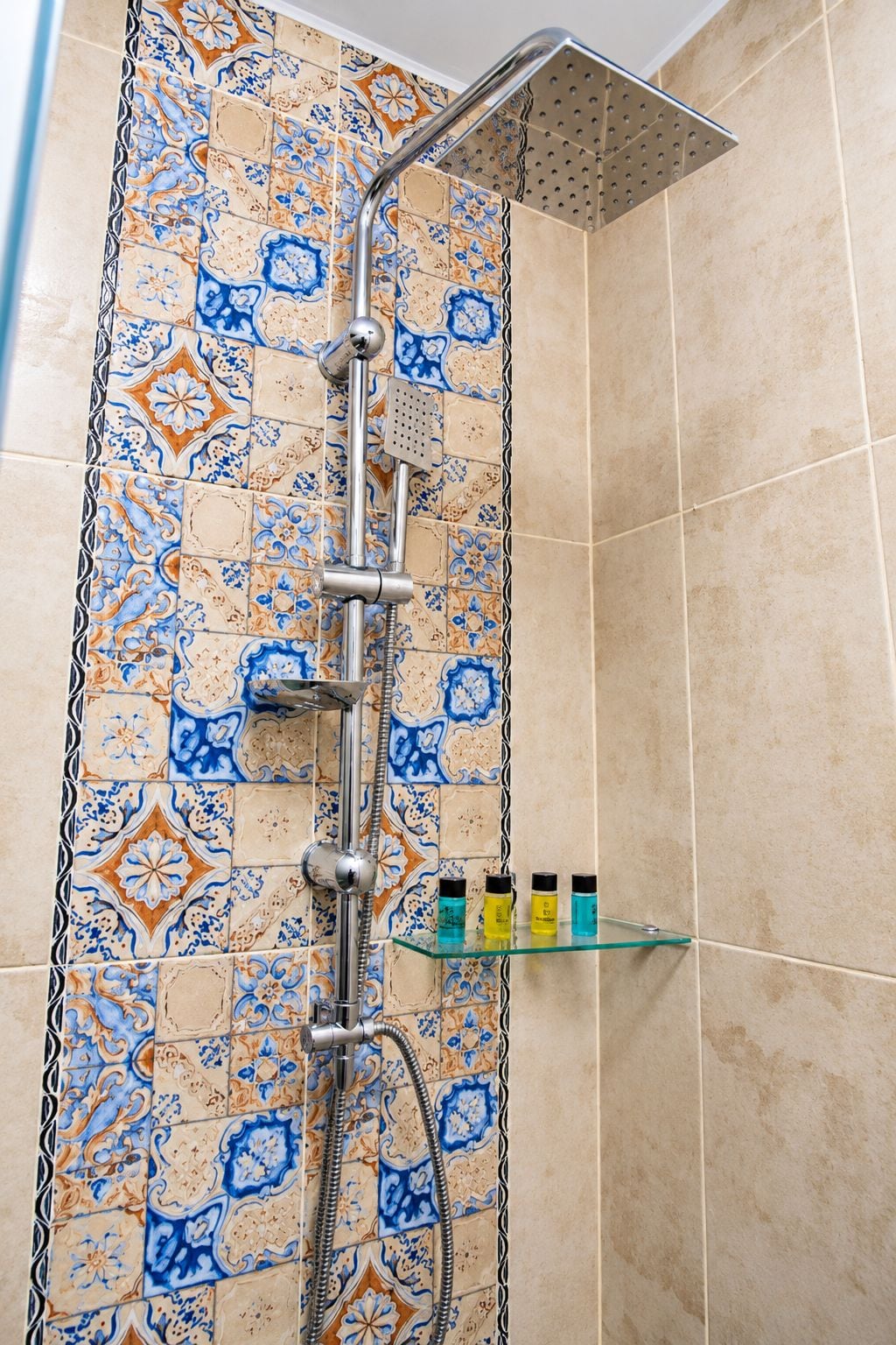 A shower with a rain showerhead and a handheld showerhead, a glass shelf with toiletries, and a tiled wall with a blue and orange pattern.