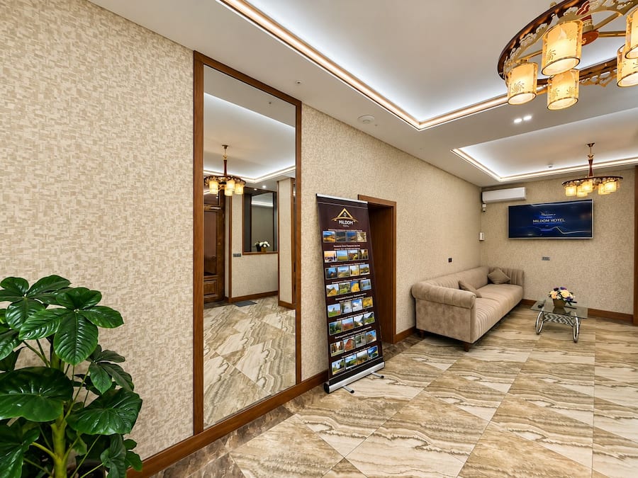 A hotel lobby with a large mirror, a reception desk, a sofa, a TV screen, and a plant.