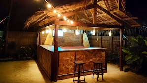 A wooden tiki bar with a thatched roof, string lights, and two stools. - Siargao Tropic Hostel (General Luna)