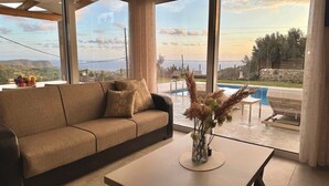 En moderne stue med sofa, sofabord og udsigt til swimmingpool og bakker.