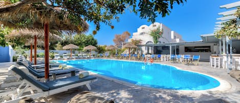 A swimming pool with lounge chairs and umbrellas, surrounded by trees and a building in the background.