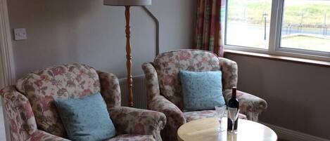 A room with two floral armchairs, a round wooden table, a floor lamp, and a window with curtains.