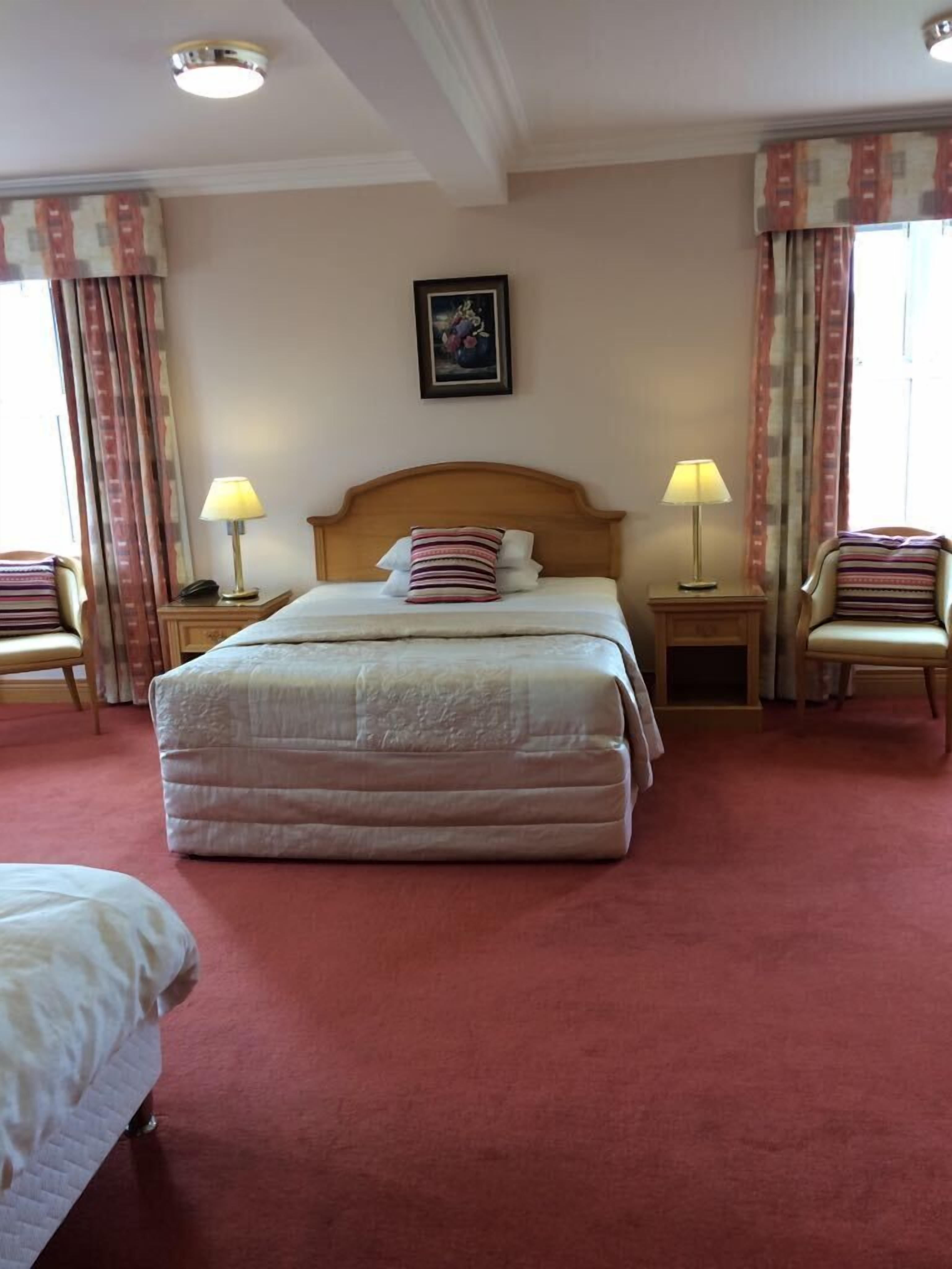 Habitación de hotel con una cama grande, dos mesitas de noche con lámparas, una silla e imagen enmarcada en la pared.