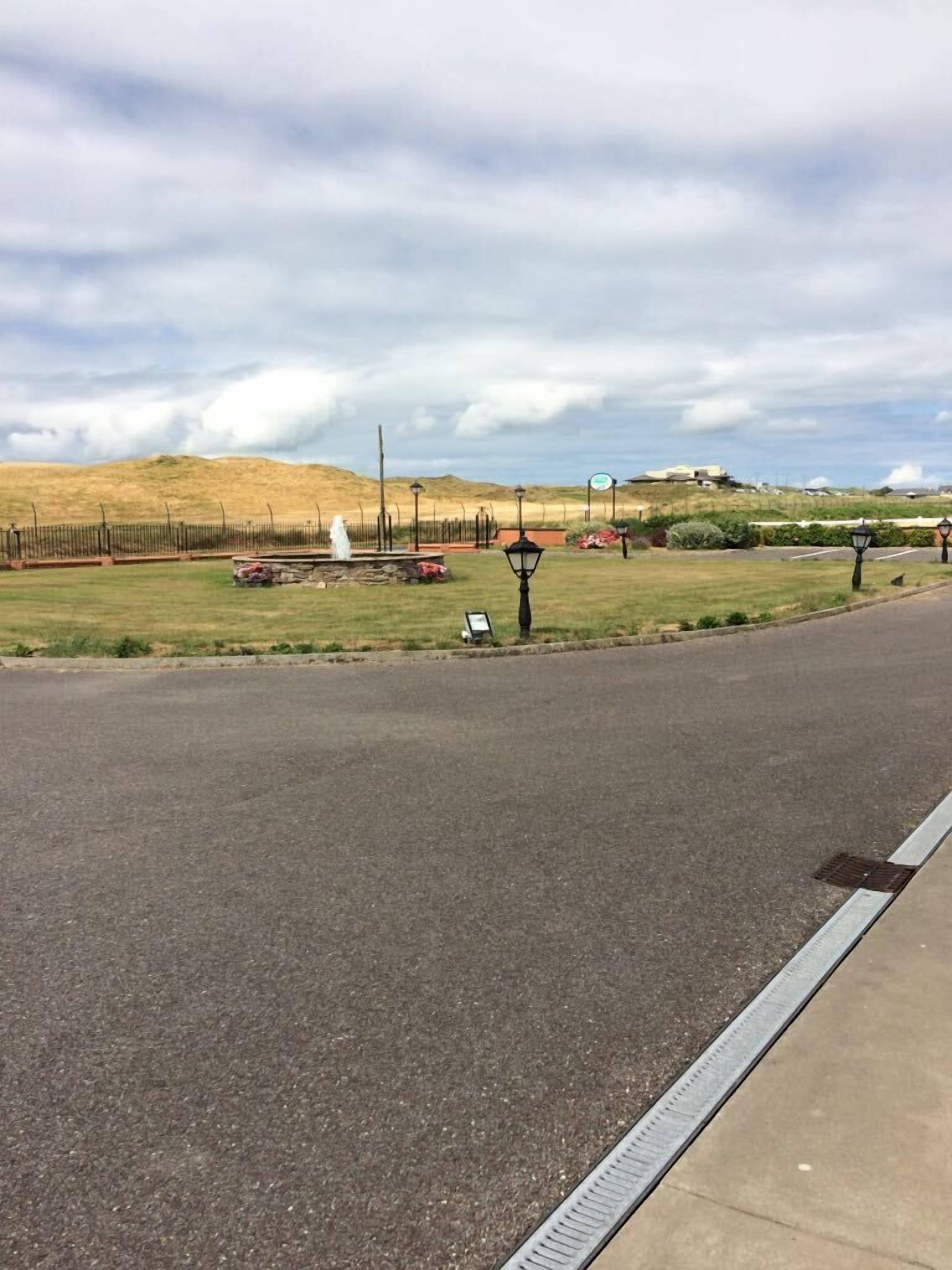 A paved road with a sidewalk, a grassy area with a statue, and a hilly landscape in the background.