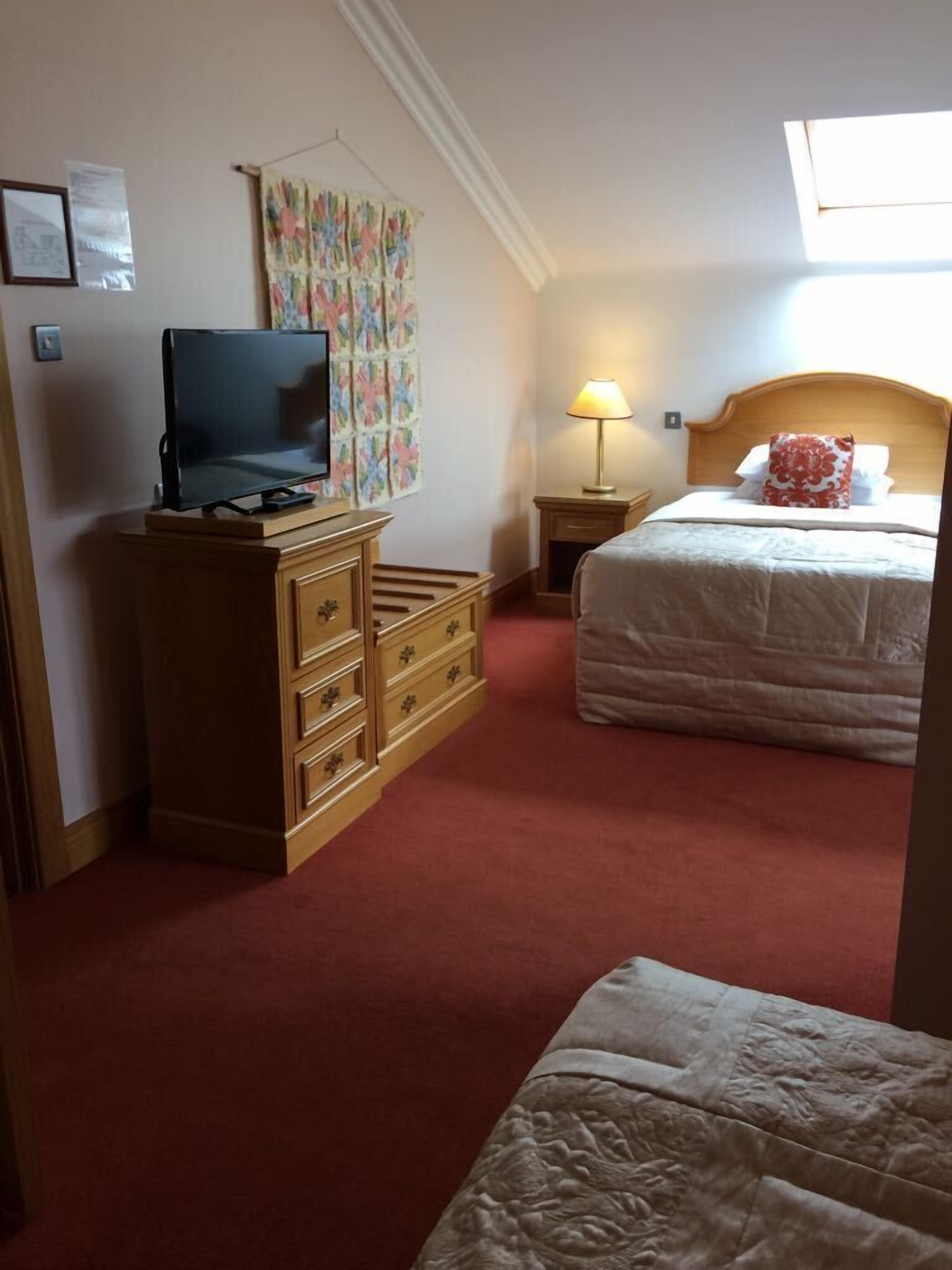 A hotel room with a TV, a wooden dresser, a bed with a red and white pillow, and a wall with a colorful artwork.