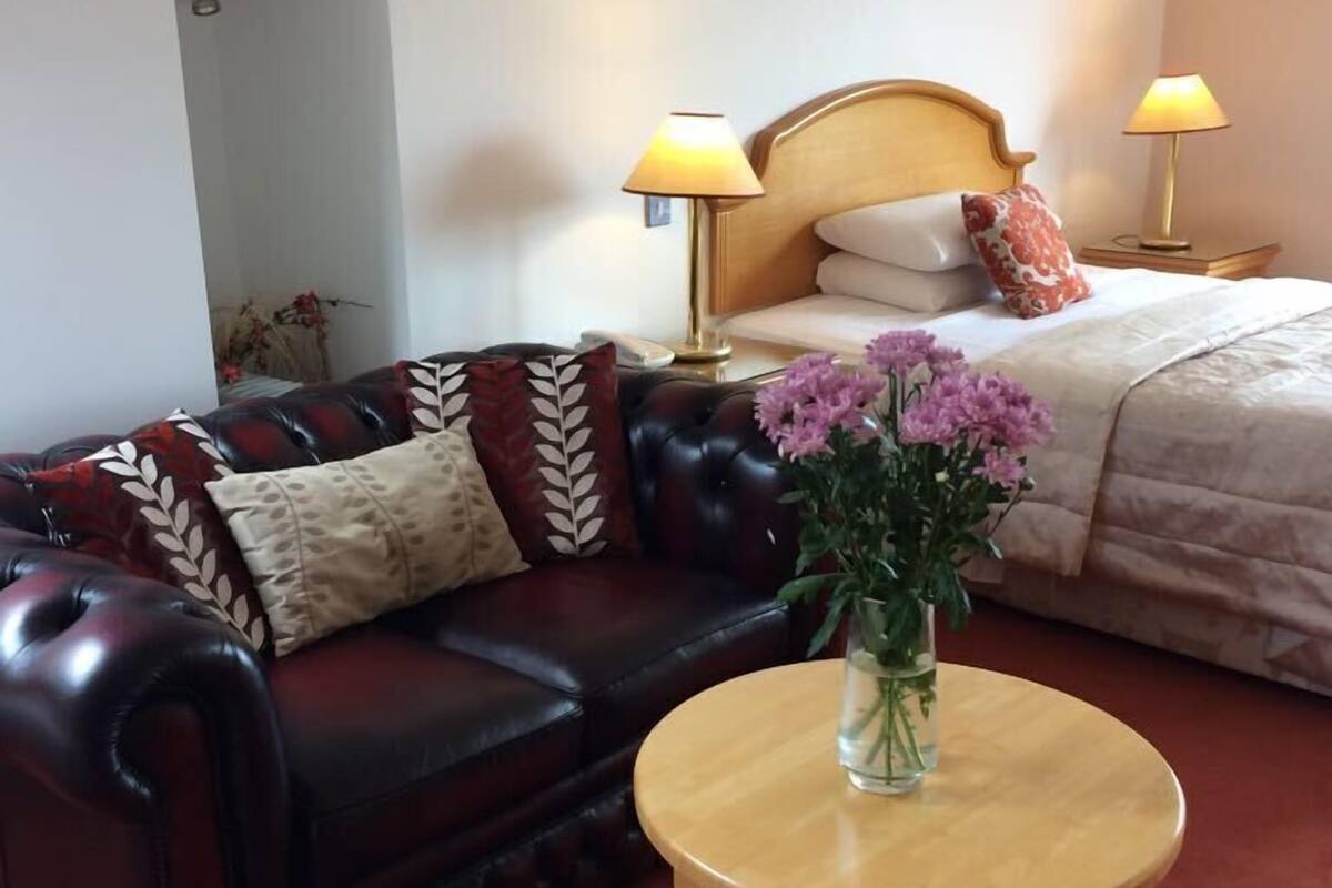 Habitación de hotel con sofá de cuero, una mesa redonda de madera, cama con dos lámparas de noche y un jarrón de flores.